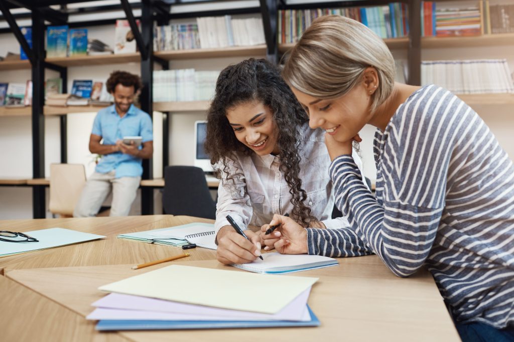 close up pair beautiful young multi ethnic student girls doing homework together writing essay presentation preparing exams with good mood (1)